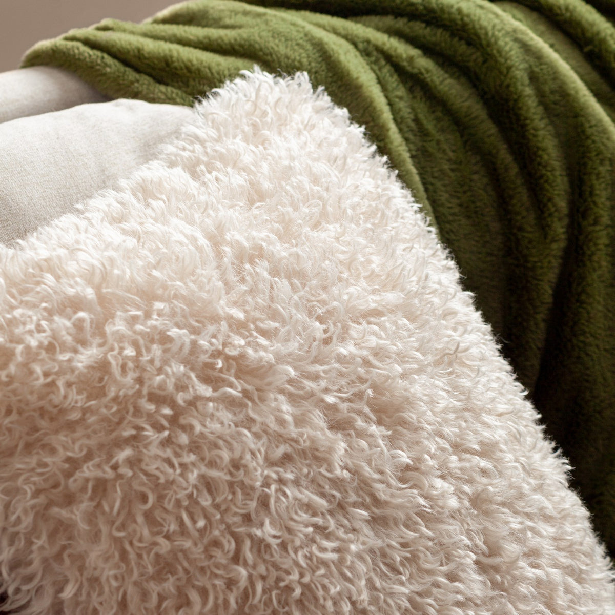 Rowan Faux Fur Polyester Velvet Cushion in natural cream, close-up of shaggy curly-pile front against a green fleece throw and beige upholstery.