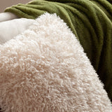 Rowan Faux Fur Polyester Velvet Cushion in natural cream, close-up of shaggy curly-pile front against a green fleece throw and beige upholstery.
