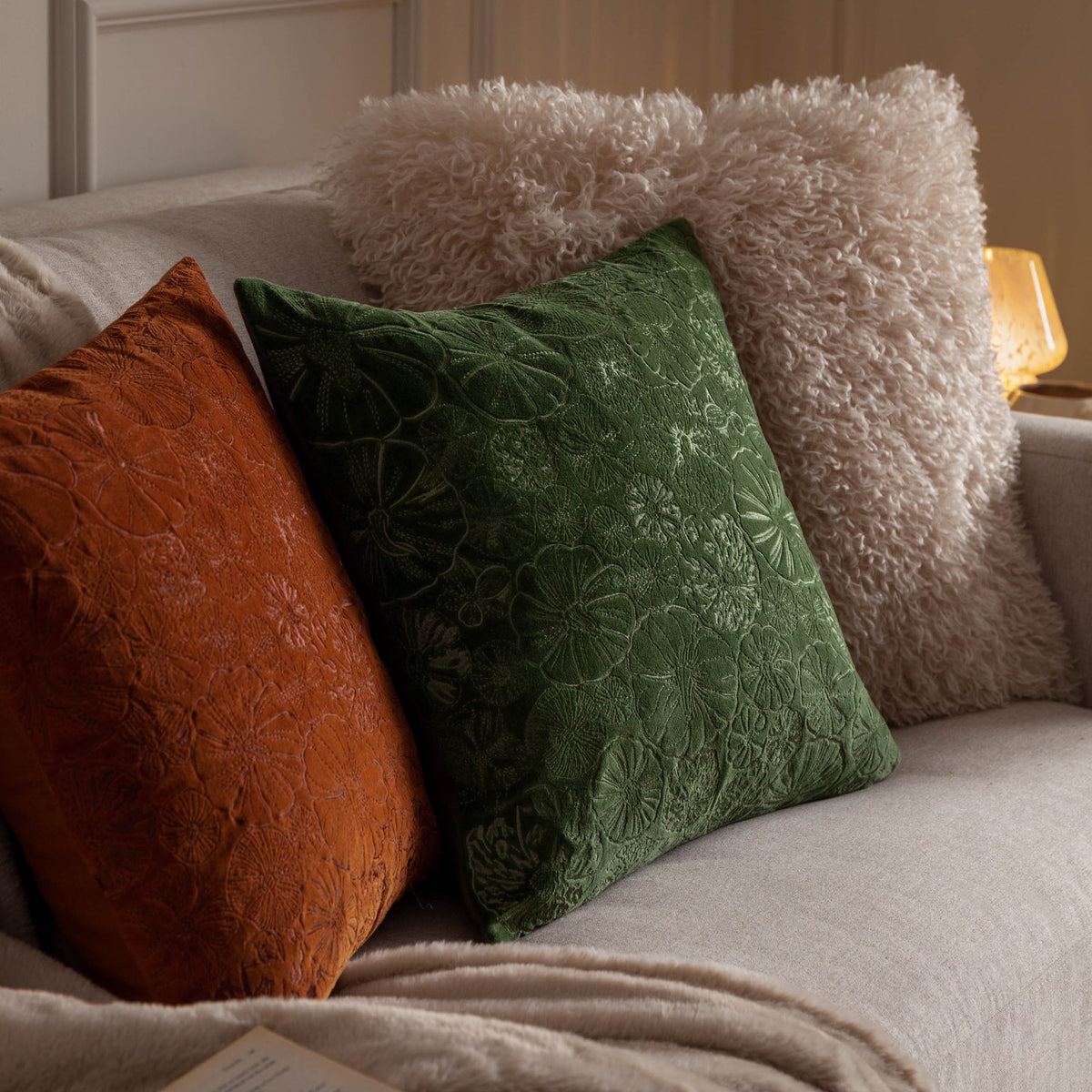 Rowan Faux Fur Cushion in natural cream with curly shaggy pile, shown on a sofa alongside rust and green velvet cushions with embroidered floral texture.