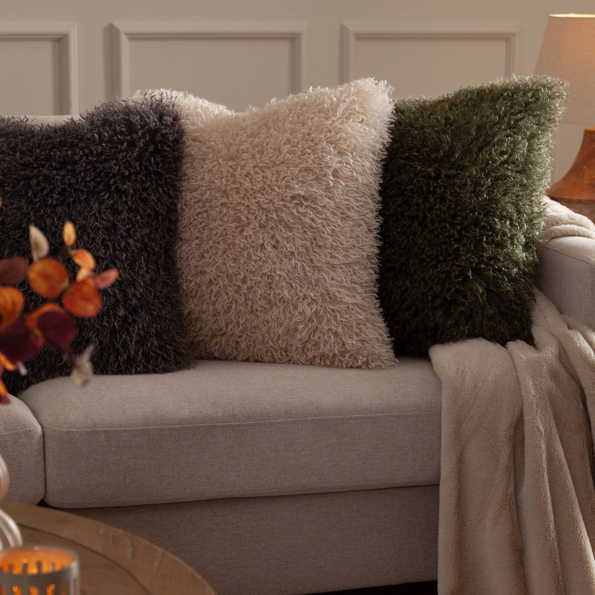 Rowan Faux Fur Polyester Velvet Cushion in natural cream with curly shaggy pile, shown centred on a beige sofa between charcoal and green faux fur cushions.