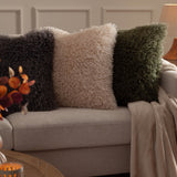 Rowan Faux Fur Polyester Velvet Cushion in natural cream with curly shaggy pile, shown centred on a beige sofa between charcoal and green faux fur cushions.