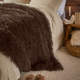 Rowan Velvet Faux Fur Throw in chocolate brown, shaggy long-pile texture draped over a bed with cream linens in a bedroom setting.