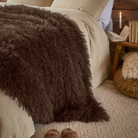 Rowan Velvet Faux Fur Throw in chocolate brown, shaggy long-pile texture draped over a bed with cream linens in a bedroom setting.