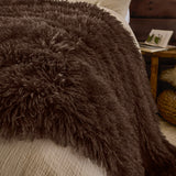 Rowan Velvet Faux Fur Throw in chocolate brown, shown draped over a bed with long shaggy pile, styled in a bedroom beside a wooden side table.