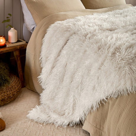 Rowan Faux Fur Plain Velvet Throw in ecru draped over a beige bed, showing its shaggy Mongolian-style fur texture in a bedroom setting.