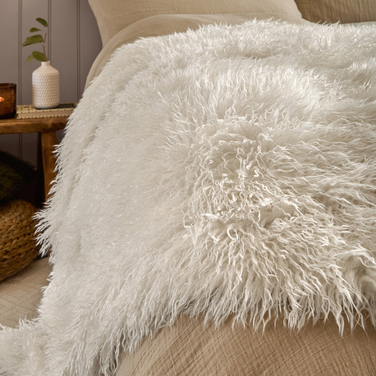 Rowan Faux Fur Plain Velvet Throw in ecru, shaggy Mongolian-style fur texture draped over a bed beside cream pillows in a bedroom setting.