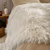 Rowan Faux Fur Plain Velvet Throw in ecru, shaggy Mongolian-style fur texture draped over a bed beside cream pillows in a bedroom setting.