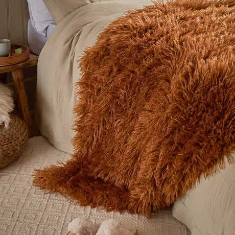 Rowan Faux Fur Plain Velvet Throw in ginger, shaggy Mongolian-style fur fabric draped over a bed with linen bedding in a bedroom setting.