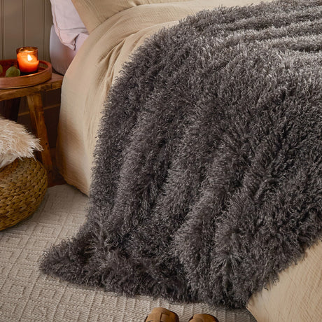 Rowan Faux Fur Plain Velvet Throw in Grey draped over a bed, showing its curly Mongolian-style fur texture in mottled charcoal with silvery tips, styled with cream bedding and a woven side stool.