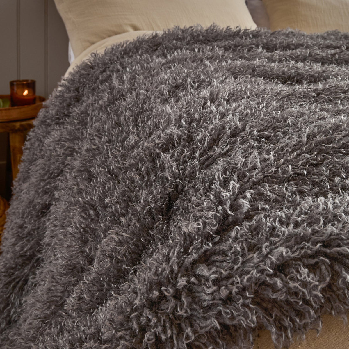 Rowan Faux Fur Plain Velvet Throw in Grey, curly Mongolian-style fur with silvery tips draped over a bed with cream pillows in a bedroom setting.