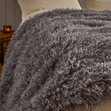 Rowan Faux Fur Plain Velvet Throw in Grey, curly Mongolian-style fur with silvery tips draped over a bed with cream pillows in a bedroom setting.