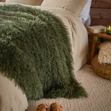 Rowan Faux Fur Plain Velvet Throw in khaki draped over a bed, showing its shaggy Mongolian-style fur texture against neutral bedding in a rustic bedroom setting.