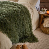 Rowan Faux Fur Plain Velvet Throw in khaki draped over a bed with shaggy fur texture, styled in a bedroom with cream bedding and a wooden bedside table.