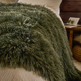 Rowan Faux Fur Plain Velvet Throw in khaki with long shaggy fur texture, draped over a bed beside cream bedding in a bedroom setting.
