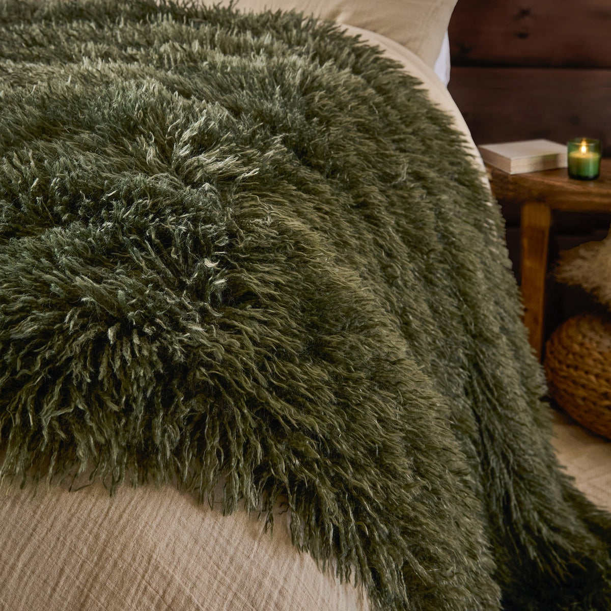 Rowan Faux Fur Plain Velvet Throw in khaki draped over a bed, showing its long shaggy fur texture beside neutral bedding, a wooden side table and candle.