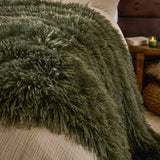 Rowan Faux Fur Plain Velvet Throw in khaki draped over a bed, showing its long shaggy fur texture beside neutral bedding, a wooden side table and candle.