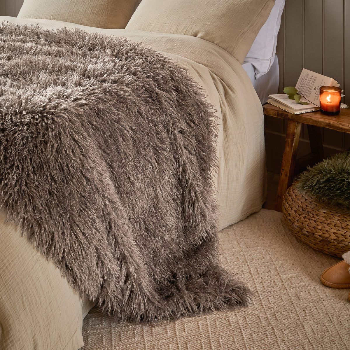 Rowan Faux Fur Plain Velvet Throw in mocha, shown draped over a bed with long-pile shaggy fur texture, beside a wooden side table with a candle in a bedroom setting.