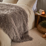 Rowan Faux Fur Plain Velvet Throw in mocha, shown draped over a bed with long-pile shaggy fur texture, beside a wooden side table with a candle in a bedroom setting.