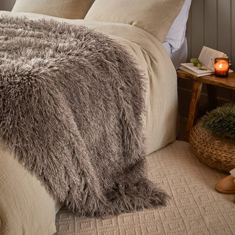 Rowan Faux Fur Plain Velvet Throw in mocha, shown draped over a bed with long-pile shaggy fur texture, beside a wooden side table with a candle in a bedroom setting.