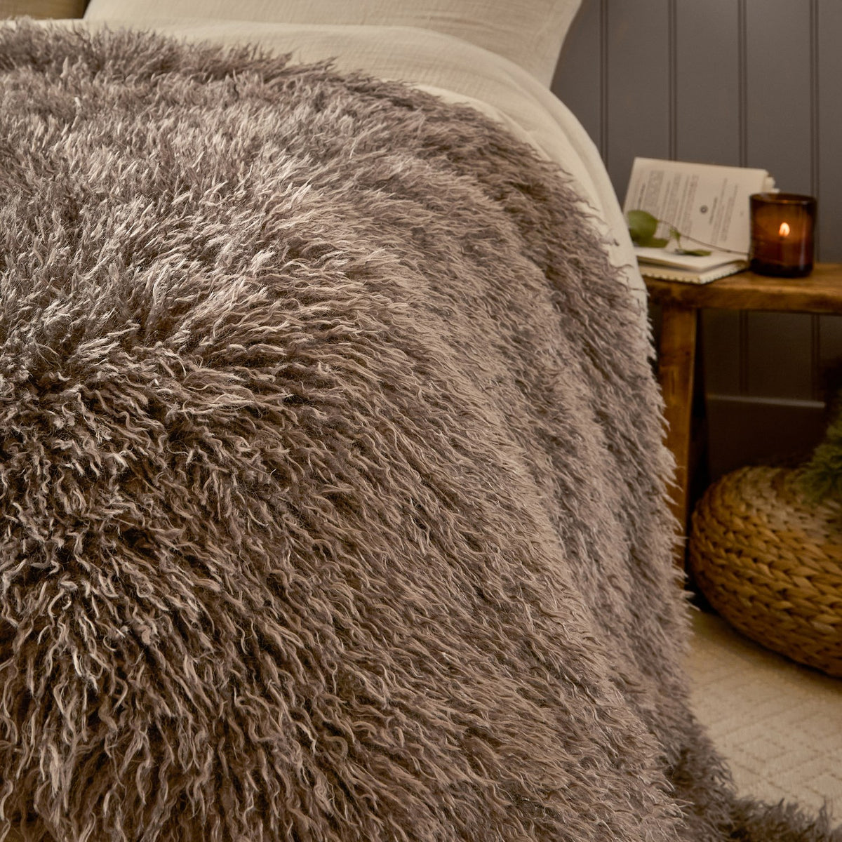 Rowan Faux Fur Plain Velvet Throw in mocha, shown draped over a bed with long shaggy fur texture, styled beside a wooden nightstand with a candle and book.