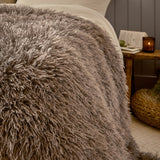 Rowan Faux Fur Plain Velvet Throw in mocha, shown draped over a bed with long shaggy fur texture, styled beside a wooden nightstand with a candle and book.