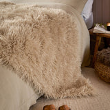 Rowan Faux Fur Plain Velvet Throw in natural beige, shaggy Mongolian-style fur draped over a bed with linen bedding, styled beside a wooden side table.