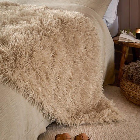 Rowan Faux Fur Plain Velvet Throw in natural beige, shaggy Mongolian-style fur draped over a bed with linen bedding, styled beside a wooden side table.