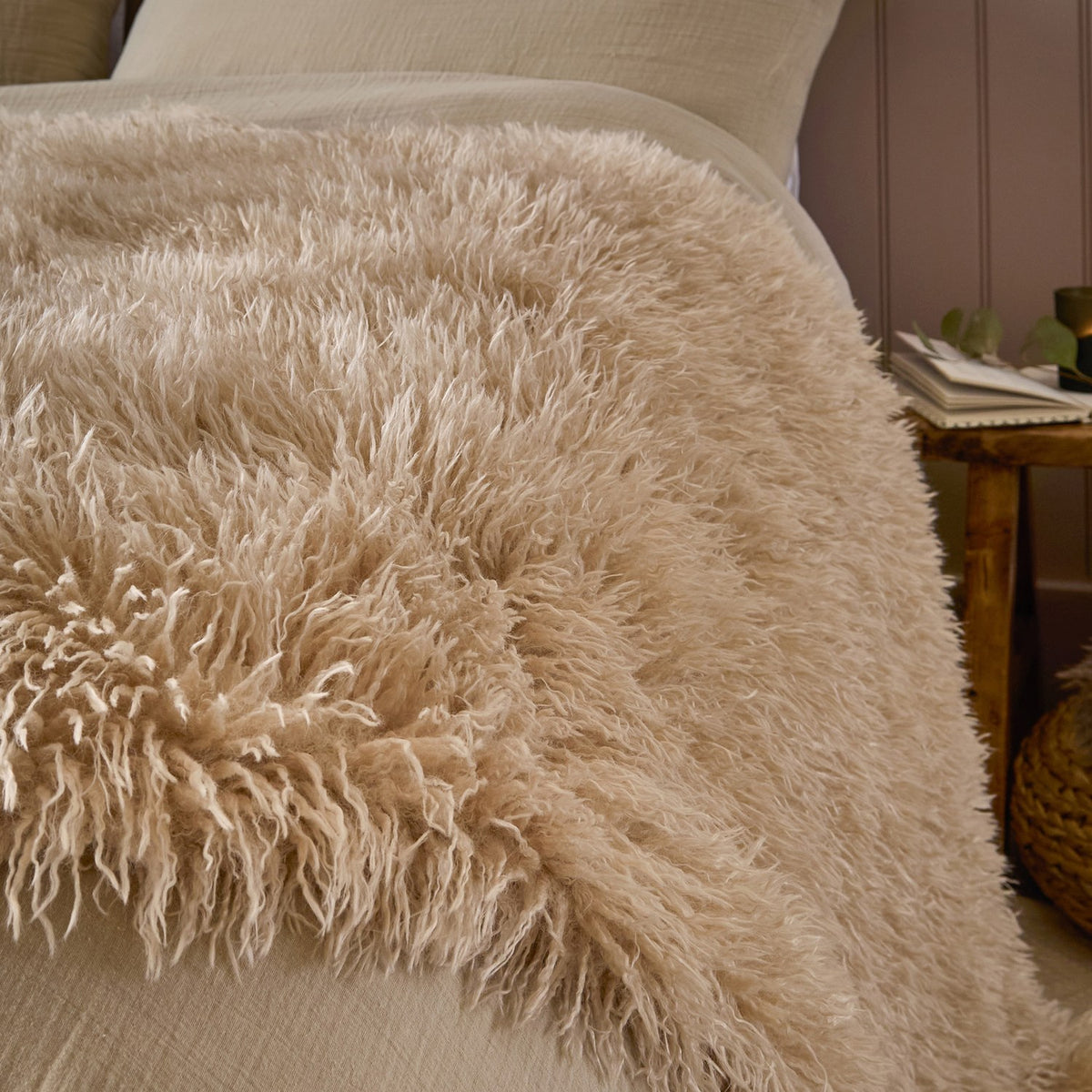 Rowan Faux Fur Plain Velvet Throw in natural beige, shown draped over a bed with its shaggy Mongolian-style fur texture visible, in a bedroom setting.