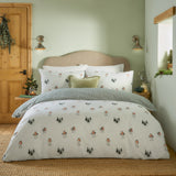 Robins Return cotton duvet cover set in cream with a robin and fir tree print, reverse side in green and cream stripe, styled on a bed in a sage green bedroom.