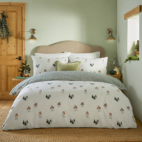 Robins Return cotton duvet cover set in cream with a robin and fir tree print, reverse side in green and cream stripe, styled on a bed in a sage green bedroom.