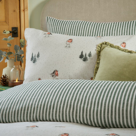 Robins Return cotton duvet cover set in cream with robin and fir tree print, styled on a bed with its reverse green and cream striped pillowcases and a green cushion.