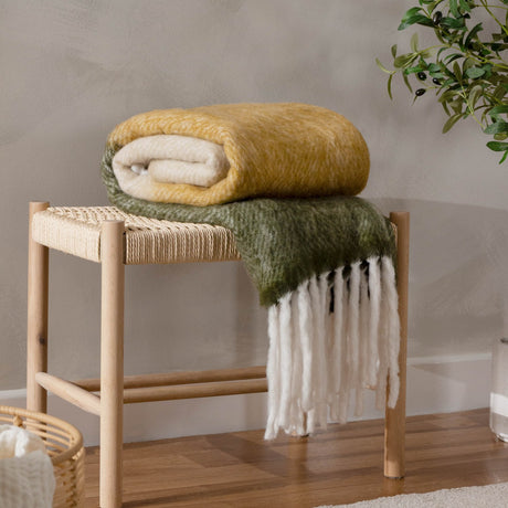 Rawton Ombre Herringbone Wool Throw in Natural/Moss, folded on a wooden bench with fringed edges trailing, ombre shading from cream to moss green, in a living room setting.