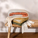 Rawton Ombre Herringbone Wool Throw in Natural/Moss, folded with fringed ends, stacked with orange and mustard throws on a wooden bench against a light wall.