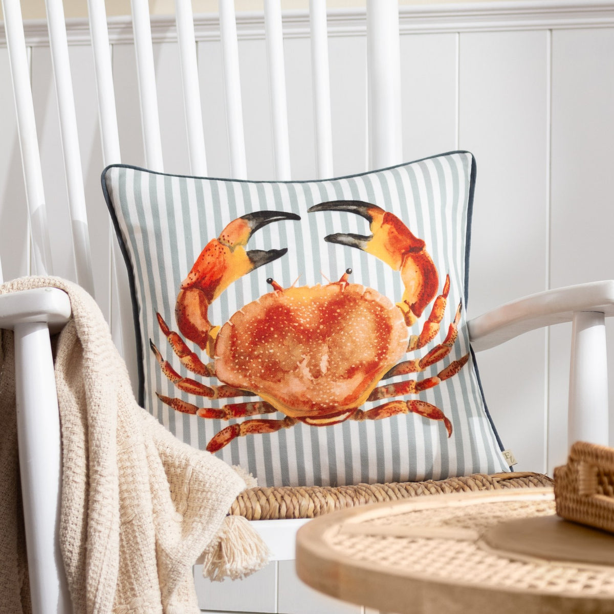 Salcombe Crab velvet cushion in orange and rust tones on a grey-blue and white striped ground, shown on a white rocking chair with a knitted throw.