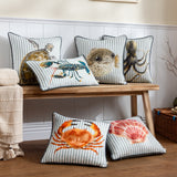 Salcombe Crab Animal Velvet Cushion in orange and rust on a grey-blue and white striped ground, shown among coordinating coastal-themed cushions on a wooden bench.
