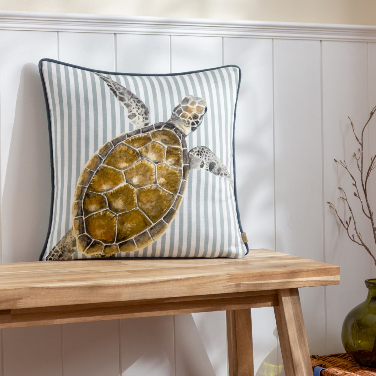Salcombe Turtle cushion with a watercolour-style turtle print in brown and amber tones on a blue-and-white striped fabric, dark piped edge, shown on a wooden bench against panelled wall.