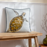 Salcombe Turtle cushion with a watercolour-style turtle print in brown and amber tones on a blue-and-white striped fabric, dark piped edge, shown on a wooden bench against panelled wall.