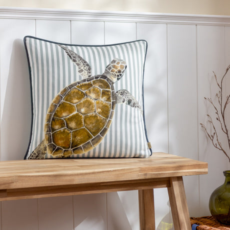 Salcombe Turtle cushion with a watercolour-style turtle print in brown and amber tones on a blue-and-white striped fabric, dark piped edge, shown on a wooden bench against panelled wall.