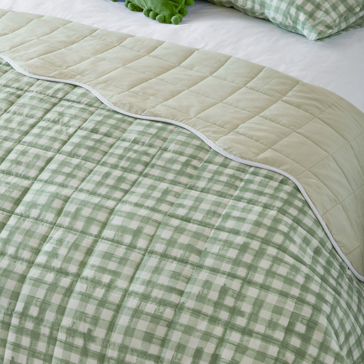 Reversible Scallop Plain Cotton Blend Bedspread in Sage, showing quilted plain sage side and gingham check reverse with white scalloped piped trim on a bed.