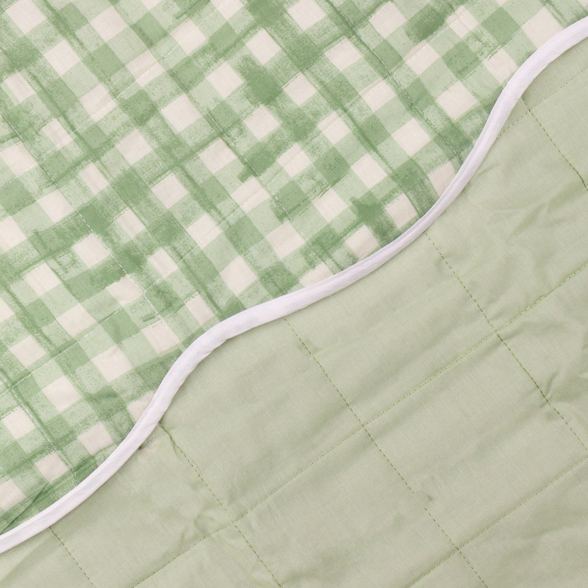 Close-up of the Scallop Plain Cotton Blend Bedspread showing its reversible design, with a sage green gingham check quilted side meeting a plain sage green quilted side, edged in white scalloped trim.