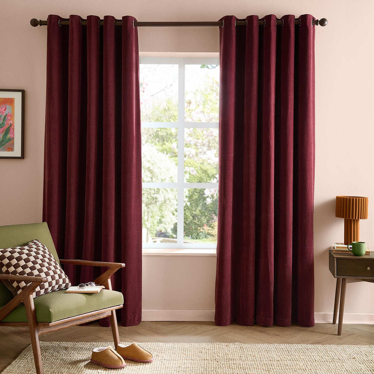 Soft Chenille Plain Lined Eyelet Curtains in deep beetroot red, hung on a dark pole either side of a window in a living room with a green armchair and side table.