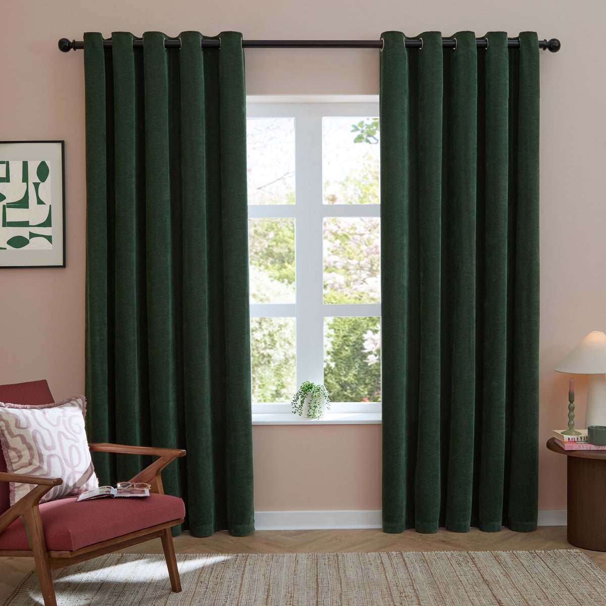 Soft chenille eyelet curtains in bottle green hang floor-length either side of a window with black rod, styled in a living room with pink walls and armchair.