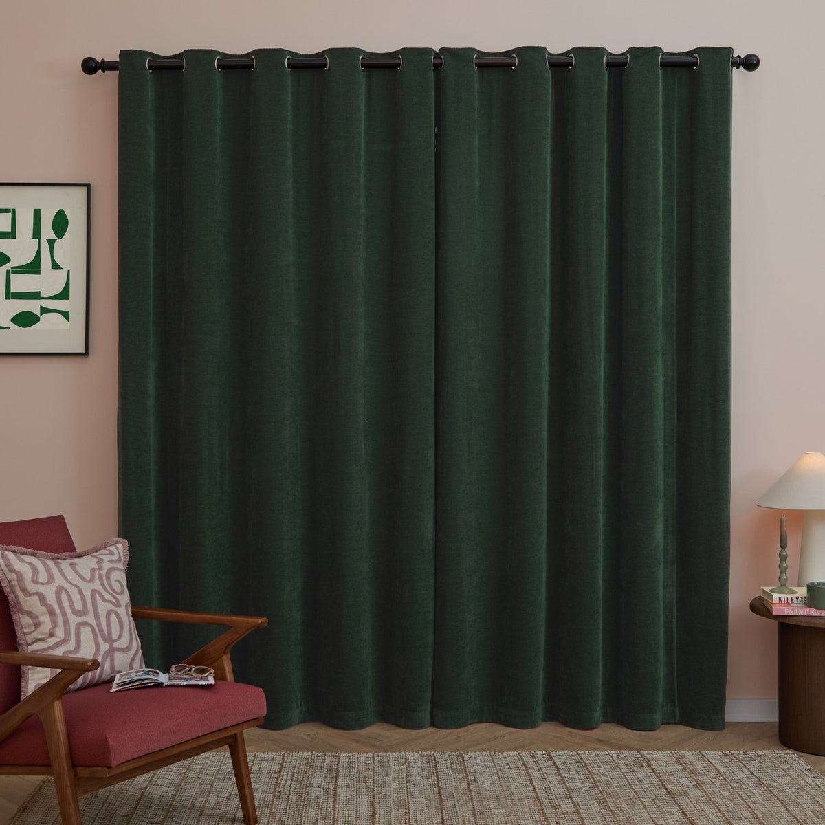 Bottle green eyelet curtains in soft chenille fabric hang full-length with dark grommets on a black rod, shown in a living room with pink walls.