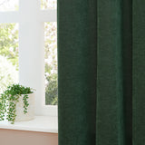 Bottle green chenille eyelet curtain hanging in folds beside a white-framed window with a potted plant on the sill.