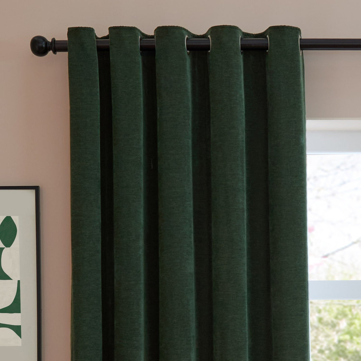 Soft Chenille Plain Lined Eyelet Curtains in bottle green hang from a black eyelet rod, showing a velvety woven texture in a bedroom setting with a window and framed print nearby.