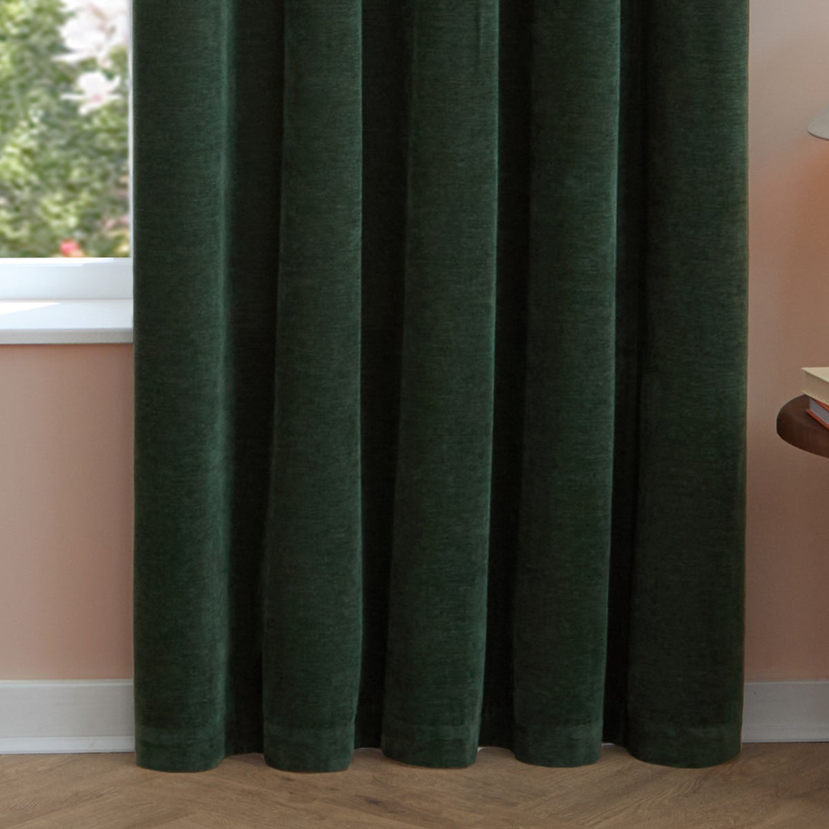 Soft chenille eyelet curtains in bottle green hanging in soft folds beside a window, shown in a bedroom with pink walls and wooden flooring.