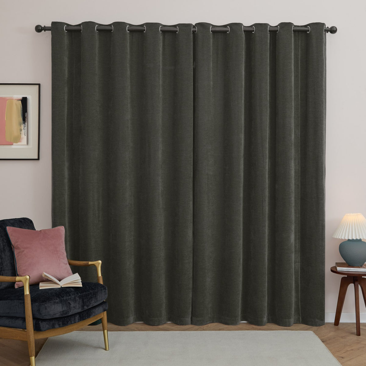 Charcoal grey chenille eyelet curtains hung on a black rod in a living room, shown with a velvet armchair, side table, and framed wall art.