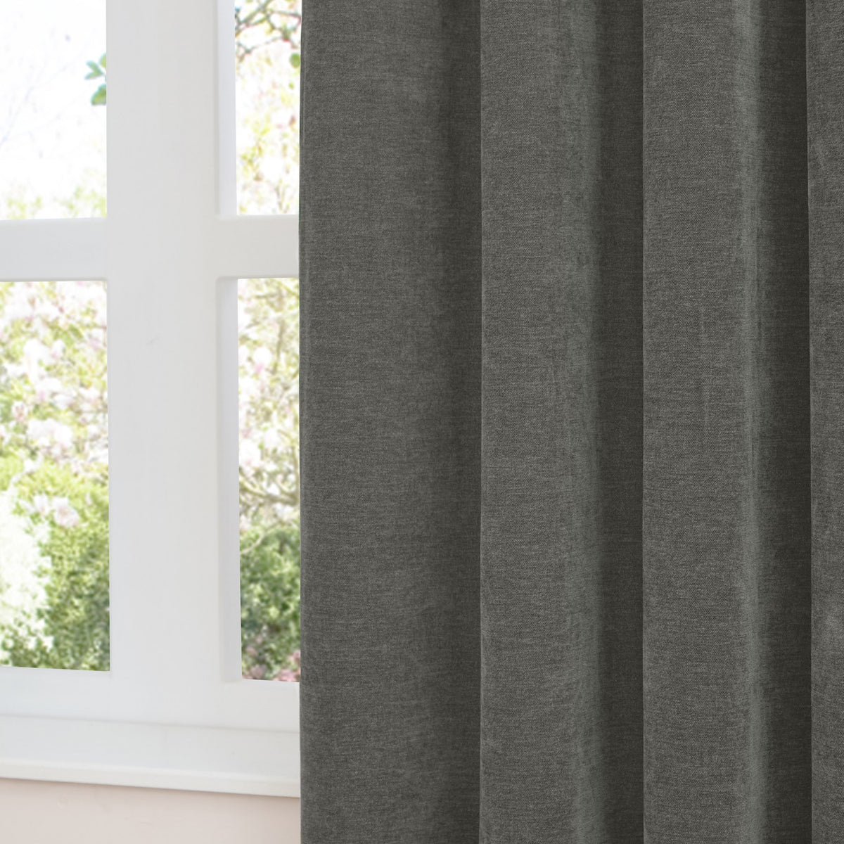 Charcoal grey chenille eyelet curtain in soft folds by a white-framed window, showing dense matt-textured fabric with a garden view beyond.