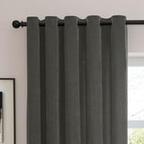Charcoal grey eyelet curtains in soft chenille fabric, shown hanging on a black rod against a light wall in a room setting.