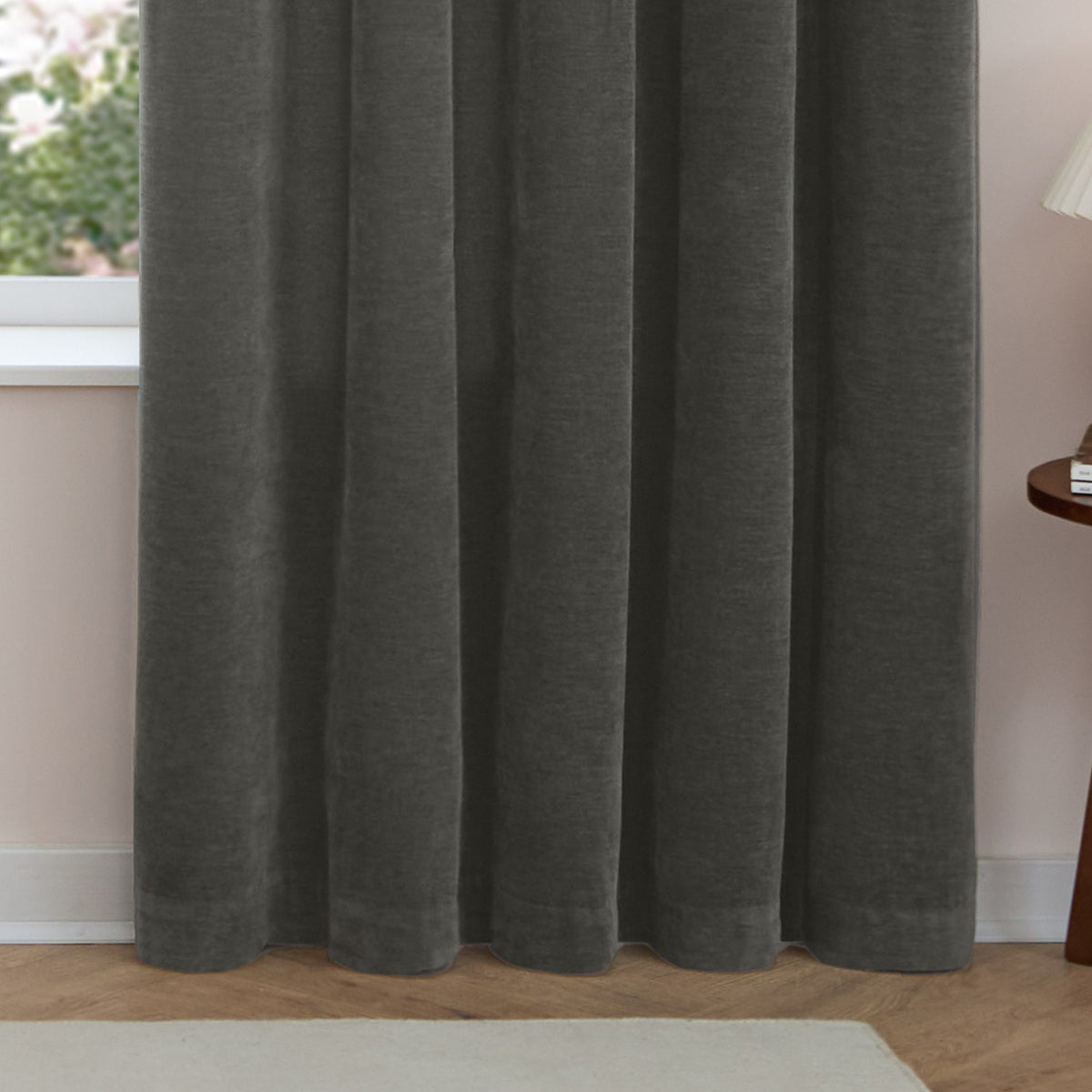 Soft chenille eyelet curtains in charcoal grey, shown falling in soft folds from ceiling to floor in a living room with pale pink walls and wooden flooring.